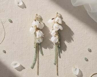 May include: A pair of elegant drop earrings featuring clusters of white fabric flowers. The earrings have gold-colored accents and strands of green beads and gold chains, creating a delicate and feminine design.