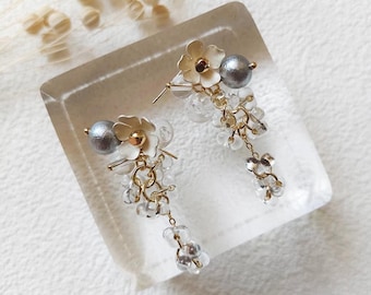 May include: A pair of dangle earrings featuring cream-colored flower charms, metallic grey spherical beads, and clusters of small clear glass beads suspended from gold-toned metal chains. The earrings are designed with a post-back closure mechanism.