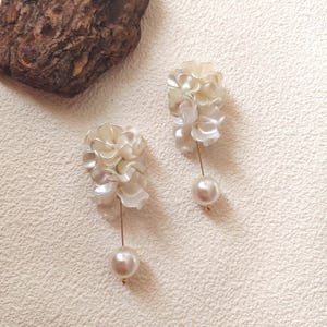 May include: A pair of elegant, off-white floral earrings. Each earring features a cluster of layered, petal-like shapes and a dangling, pearlescent bead. The earrings are connected by a thin, gold-colored post. The earrings are set against a textured, light-colored background.
