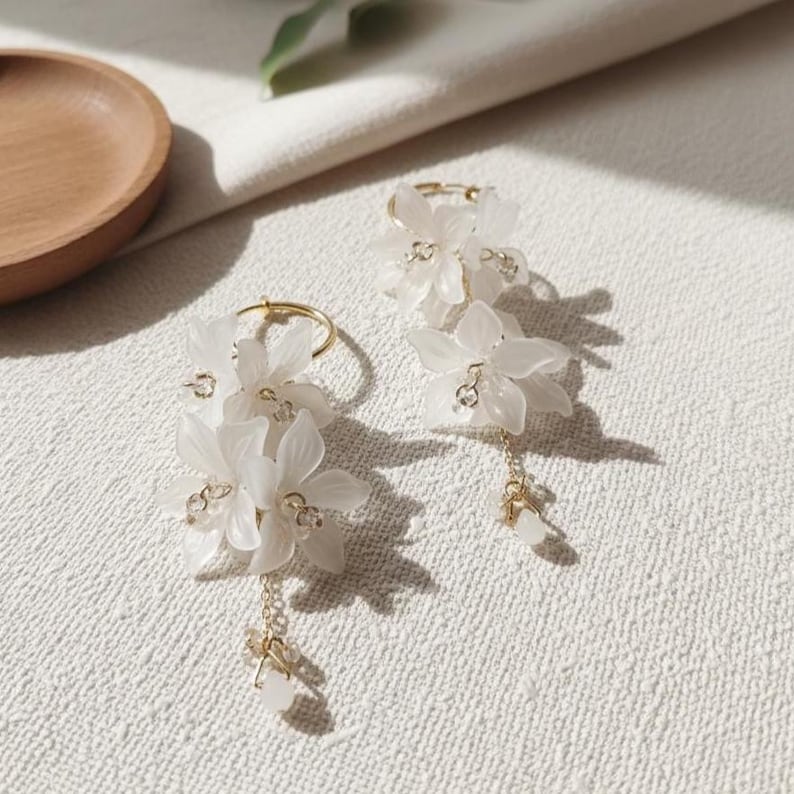 May include: A pair of delicate white flower earrings. Each earring features a gold hoop with cascading white flower petals and small, sparkling accents. The earrings dangle with a small cluster of white beads.