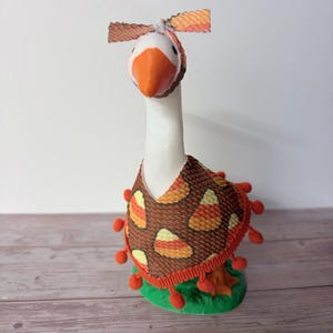 May include: A white goose figurine wearing a brown cape with a candy corn pattern and orange pom-pom trim. The goose has an orange beak and a matching brown and orange candy corn patterned headband with a bow on top, standing on a green base.