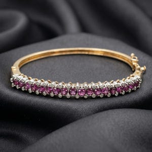 Può includere: Un bracciale rigido dorato con una fila di pietre preziose rosa e piccole pietre bianche. Il bracciale è posto su uno sfondo nero e setoso, che mette in risalto la lucentezza e il design del gioiello.
