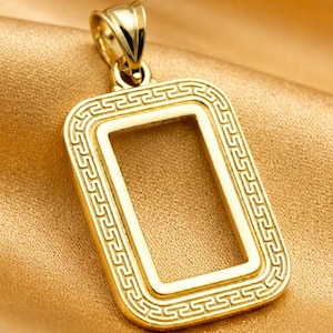 Puede incluir: Un colgante rectangular dorado con un centro abierto y un borde decorativo. El colgante está suspendido de un pequeño lazo y descansa sobre una tela de satén dorado. El diseño presenta un patrón repetitivo en los bordes.