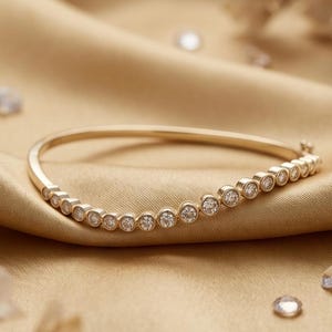 May include: A gold-colored bracelet with a row of small, round, clear gemstones. The bracelet is set against a gold-colored, silky fabric background, with scattered gemstones and floral accents.