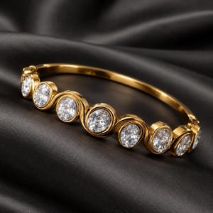 May include: A gold-toned hinged bangle bracelet featuring a series of seven round, clear, faceted stones set in individual bezels. The metal frame has a continuous, undulating wave-like design that connects each stone setting in a row.