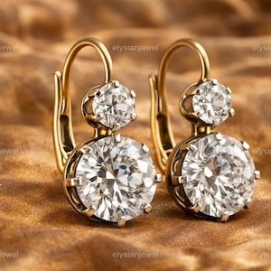 May include: A pair of gold-colored leverback earrings with two clear, round-cut gemstones. The earrings are set against a warm, golden-brown background, creating a luxurious and elegant aesthetic. The gemstones sparkle, reflecting light.