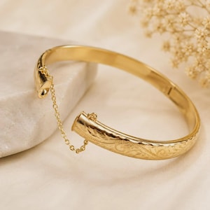 May include: A gold-colored bangle bracelet with an engraved floral pattern. The hinged bracelet has a safety chain and is displayed on a white surface. The jewelry is a classic accessory.