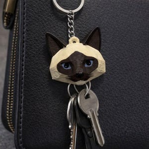 May include: A Siamese cat keyring with a silver chain and key ring. The cat's face is tan and brown with blue eyes. Several keys hang below the keyring. The background is a black leather surface.