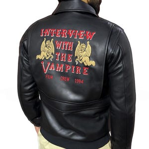 May include: Black leather jacket with the text "INTERVIEW WITH THE VAMPIRE" in red and gold embroidery on the back. The jacket features a classic design with a collar and a tailored fit. The text includes the year "1994".