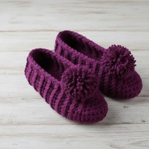 Pode incluir: Um par de pantufas de crochê roxas feitas à mão. As pantufas têm uma textura canelada e um detalhe de pompom na frente. As pantufas são projetadas para uso interno e são feitas de fio macio.