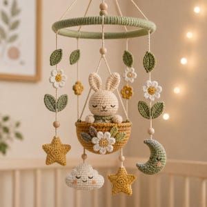 Peut inclure: Un mobile pour bébé en crochet avec un lapin beige central assis dans un panier jaune moutarde. Le mobile comprend des éléments suspendus tels que des fleurs blanches, des feuilles vertes, des étoiles jaunes, un croissant de lune vert et un nuage blanc, le tout suspendu à un anneau vert sauge.