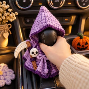 Könnte beinhalten: Ein violetter, gehäkelter Schaltknaufbezug in Form eines Kapuzenumhangs mit einem kleinen weißen Totenkopf-Akzent und einem braun-weißen Sensen-Accessoire. Der Bezug hat vorne ein Zugband und ist für die Abdeckung eines Autoschalthebels gedacht.