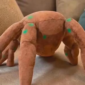 May include: A brown, multi-legged plush toy featuring a rounded central body and several elongated, segmented limbs. The surface of the brown fabric is decorated with small, bright green circular spots distributed across the legs and the main body.