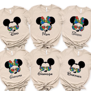 Beige family matching autism awareness shirts, featuring Mickeystyle ears, sunglasses or bows with colorful autism puzzle pieces, labeled Dad, Mom, Sister, Brother, Grandma, and Grandpa.
