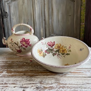 Puede incluir: Dos piezas de cerámica antiguas: un jarrón con asa y un cuenco grande. Ambos son de color crema con diseños florales en tonos rosa, amarillo y verde. El jarrón tiene un asa grande y el cuenco tiene un borde decorativo.