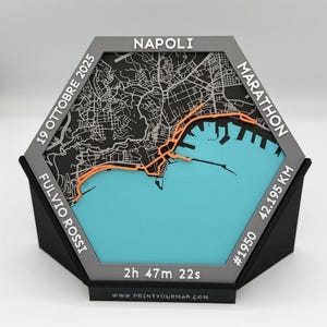 Op de afbeelding: Een zeshoekige 3D-kaart van Napels, Italië, met een oranje marathonroute. De kaart bevat de tekst "NAPOLI", "MARATHON", "19 OTTOBRE 2025", "FULVIO ROSSI", "42.195 KM" en "2h 47m 22s". De basis is zwart.
