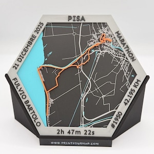 Può includere: Una mappa esagonale di Pisa, Italia, con un percorso arancione che segna un percorso di maratona. La mappa include il testo: "PISA MARATHON 42,195 KM" e "21 DICEMBRE 2025". La base è nera, con una sezione azzurra che rappresenta il mare.
