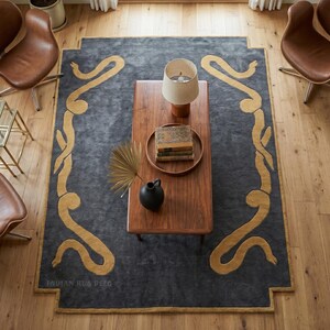 Op de afbeelding: Een rechthoekig vloerkleed met een donkergrijs veld en een mosterdgeel, slangachtig randpatroon. Het kleed ligt op een lichte houten vloer, gecentreerd onder een houten salontafel met een lamp, boeken en een zwarte vaas.