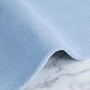 Pode incluir: Close-up de um tapete azul claro com uma textura macia e felpuda. O tapete está dobrado, revelando sua espessura e a superfície de mármore por baixo. O texto "INDIAN RUG DECO" é visível no canto superior direito.