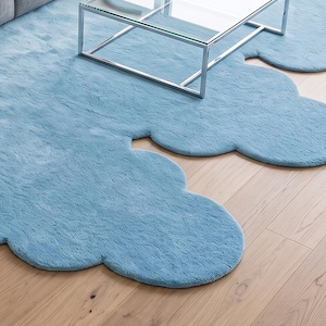 May include: A light blue area rug with a distinct cloud-like shape featuring rounded, scalloped edges and a soft, textured pile surface. The rug is positioned on a light-toned wooden floor, partially underneath a metal and glass coffee table.