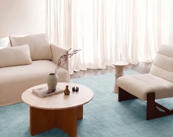 May include: A round wooden coffee table with a light-colored top and a cross-shaped base, positioned on a large light blue area rug. The table holds a small ceramic vase with dried stems, a book, a small metallic object, and a dark decorative piece.