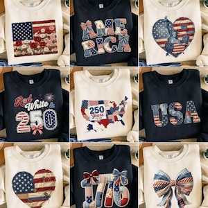 May include: A collection of cream and navy blue sweatshirts with patriotic designs. The designs include the American flag, the word "AMERICA", a heart with the Statue of Liberty, a map of the USA, the word "USA", a heart with the American flag, the number "1776", and a bow.
