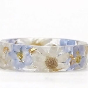 May include: A clear resin ring with embedded dried flowers and gold leaf flakes. The ring features white and light blue flowers, creating a delicate and natural aesthetic. A unique piece of jewelry.