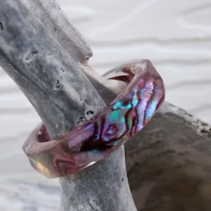 May include: A clear resin ring with iridescent abalone shell inlay. The ring features a faceted design, showcasing the vibrant blues, purples, and greens of the shell. The ring is displayed on a gray, textured surface.