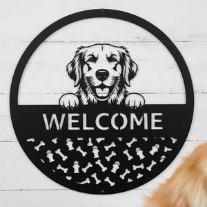 Könnte beinhalten: Ein rundes Wand-Schild aus schwarzem Metall mit einer ausgeschnittenen Illustration eines Hundes, dessen Pfoten auf einer horizontalen Leiste ruhen. Unter der Leiste ist das Wort WELCOME in Großbuchstaben ausgeschnitten, gefolgt von einem Muster aus Knochen und Hydranten.