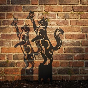 Könnte beinhalten: Ein schwarzer Gartenstecker aus Metall mit der Silhouette einer Katze, die auf den Hinterbeinen steht und nach einem Schmetterling greift. Das ausgeschnittene Design zeigt komplexe Muster im Körper und im Schwanz der Katze.