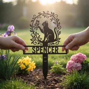 Könnte beinhalten: Ein schwarzer Metall-Gartenstecker mit der Silhouette einer Katze, die in einem blättrigen ovalen Rahmen nach einem Schmetterling greift. Unter der Katze zeigt ein horizontaler Balken den Namen SPENCER sowie die Jahreszahlen 2021 und 2026.