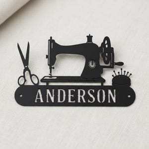 Könnte beinhalten: Schwarzes Metallschild mit einer Nähmaschine, einer Schere und einem Nadelkissen. Das Schild trägt den Namen "ANDERSON" in weißen Buchstaben, ideal für ein Bastelzimmer oder einen Nähbegeisterten.