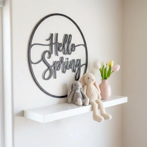 May include: A white floating shelf displays a decorative black metal sign that reads "Hello Spring." Two plush bunny rabbits and a pink vase with tulips complete the spring-themed decor. The wall is painted white.