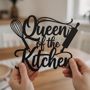 Könnte beinhalten: Schwarzes Küchenschild aus Metall mit den Worten "Queen of the Kitchen" in Schreibschrift. Das Schild zeigt einen Schneebesen und eine Teigrolle. Das Schild wird von einer Person gehalten.
