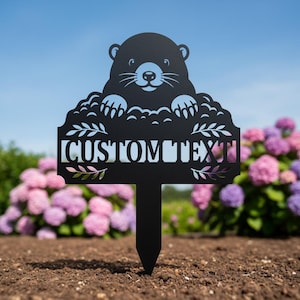 Könnte beinhalten: Schwarzer Metall-Gartenstecker mit einem Murmeltier-Design und dem Text "CUSTOM TEXT". Das Murmeltier blickt über eine dekorative Bordüre mit Blättern. Der Stecker ist in den Boden vor einem Hintergrund aus bunten Blumen gesteckt.