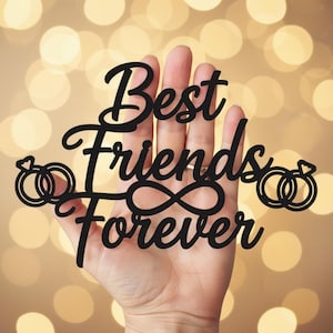 Puede incluir: Letrero recortado negro con las palabras "Best Friends Forever" en una fuente de escritura, sostenido en una mano. El diseño incluye dos iconos de anillos y un símbolo de infinito. El fondo es un bokeh suave y borroso de luces doradas cálidas.