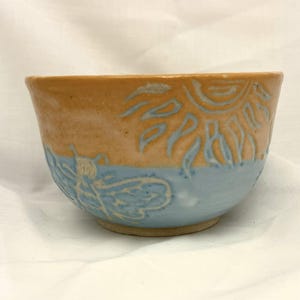 May include: A handcrafted ceramic bowl with a two-tone design. The upper section is orange, decorated with a sunburst pattern, and the lower half is light blue with a dragonfly design. The bowl has a rounded shape and a textured surface.