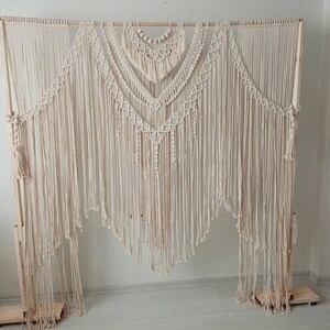 Puede incluir: Un gran tapiz de macramé de color crema. La pieza de arte textil presenta intrincados patrones de nudos, incluyendo formas de diamante y flecos. Se exhibe sobre un marco de madera, creando una estética bohemia.