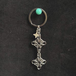 May include: A silver-coloured keyring with a decorative chainmail design. The keyring features a small star charm and a turquoise bead. The chainmail design is in the shape of a cross. The keyring is on a black background.