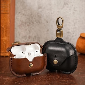 Pode incluir: Duas capas de couro para AirPods. Uma é marrom e aberta, revelando fones de ouvido brancos. A outra é preta e fechada, com um fecho cor de latão. Ambas as capas têm fecho de botão e uma estética vintage.