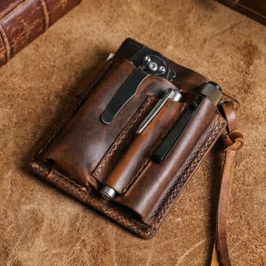 Op de afbeelding: Een bruinleren zakorganizer met meerdere compartimenten. Het bevat een vouwmes, een pen en een kleine zaklamp. De organizer heeft een gestikte rand en een leren riem met een knoop.