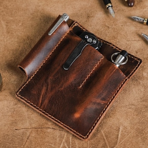 May include: A brown leather pocket organiser with a pen, a knife, and a pen with a key ring. The organiser has a rectangular shape with a stitched border. Several pens are scattered around the organiser on a brown surface.