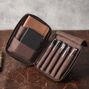 May include: A brown zippered organizer case featuring a brown notebook and a black notebook in the left pocket, and five individual pen slots on the right side containing various metallic pens with clips, plus one separate pen lying outside.