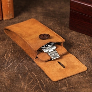 Puede incluir: Una funda para reloj de ante color canela con una solapa plegable y un cierre de botón de madera marrón oscuro. La funda contiene un reloj de pulsera de acero inoxidable con esfera negra y brazalete de eslabones metálicos, visible dentro.