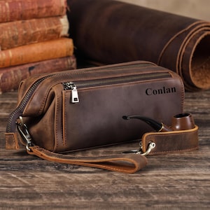Könnte beinhalten: Eine dunkelbraune Kulturbeutel aus Leder mit Reißverschluss und Handschlaufe. Der Name Conlan ist auf der Seite aufgedruckt. Eine braune Tabakpfeife ist mit einem Lederriemen und einem Metallverschluss an der Seite der Tasche befestigt.