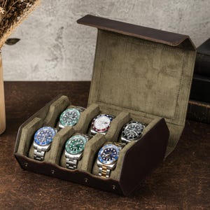 May include: A dark brown leather watch storage case with a hinged lid and an olive green velvet interior. The case features six individual compartments, each containing a stainless steel wristwatch with various colored bezels and dials.