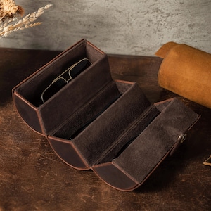 Puede incluir: Un estuche de gafas de sol de cuero marrón, abierto para revelar múltiples compartimentos forrados con tela marrón oscuro. Un par de gafas de sol descansa en el compartimento superior. El estuche está sobre una superficie marrón.