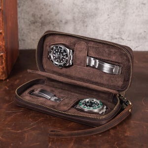 Pode incluir: Um estojo de viagem de couro castanho para relógios com fecho de correr. O estojo está aberto, revelando dois relógios e uma pulseira de relógio. Um relógio tem um mostrador preto, e o outro verde. O estojo é forrado com um material castanho macio.