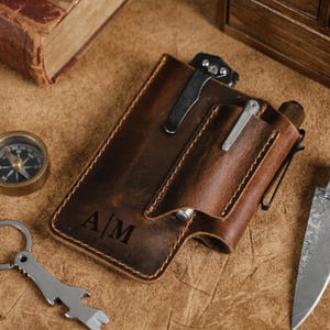 May include: A brown leather pocket organizer featuring a vertical pocket with a silver metal clip, a side loop for a pen, and the embossed initials A|M. The organizer includes a black metal pocket clip attached to the top edge.