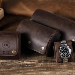 May include: Three cylindrical dark brown leather watch rolls with brass snap closures. One roll features the engraved letters FR and DEC. A silver-toned analog wristwatch with a black dial and metal link bracelet rests on a brown fabric insert.